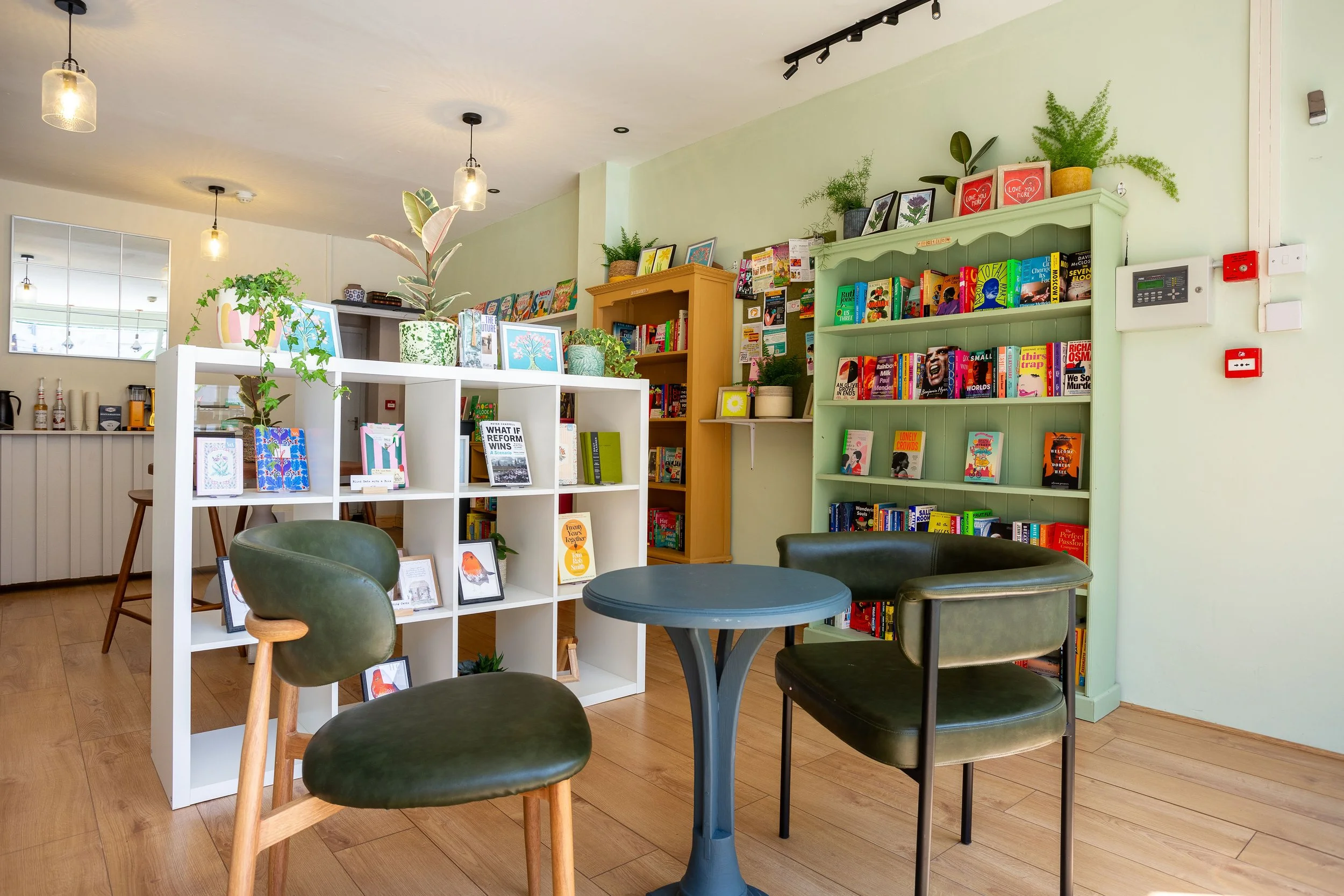 Interior of a cosy bookstore or cafe with bookshelves, a round table, chairs, and decorative plants, illuminated by warm pendant lighting.