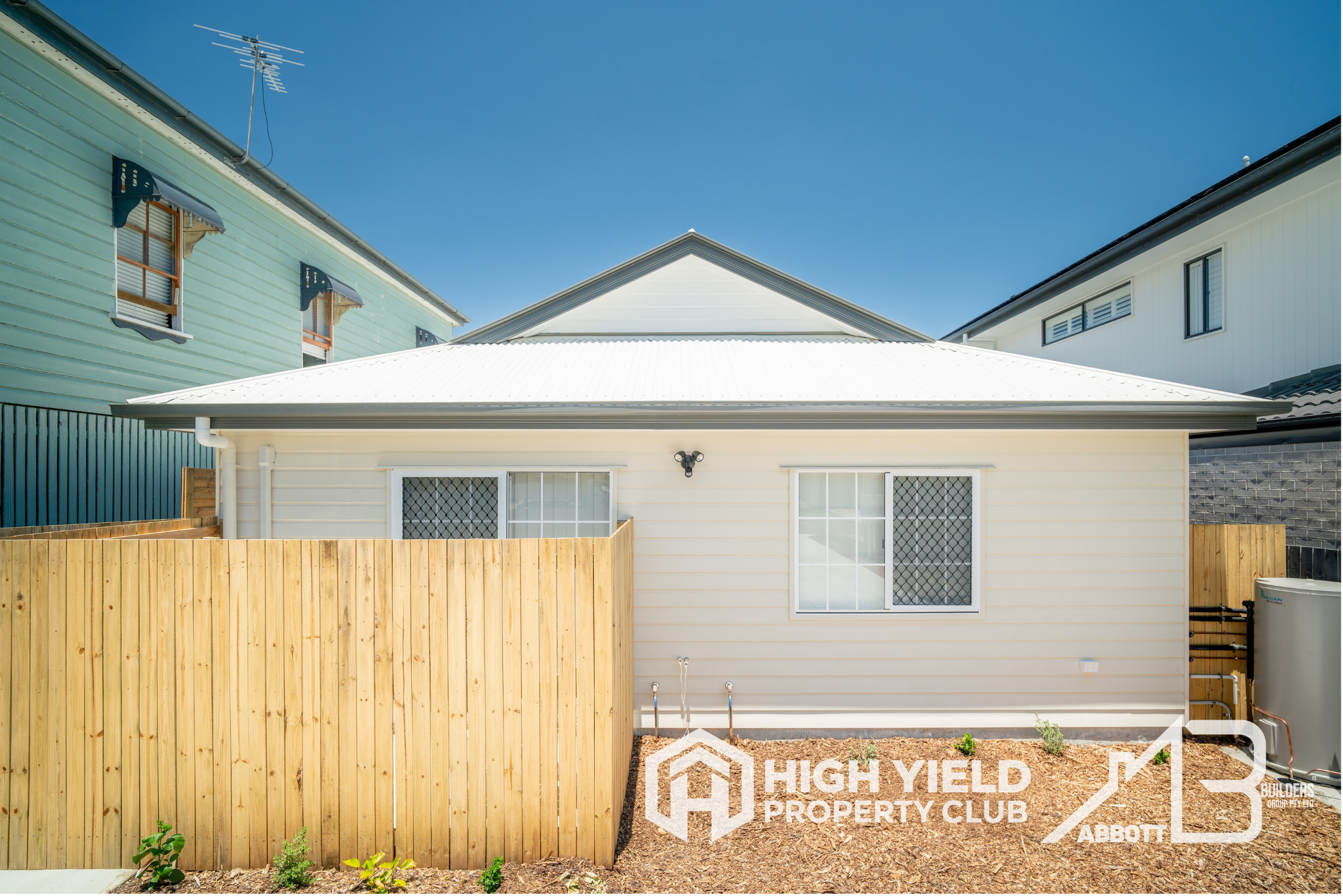 A rooming accomodation house. View of the backyard of a house with a wooden privacy fence, white house exterior, and garden bed with plants, under a clear blue sky.
