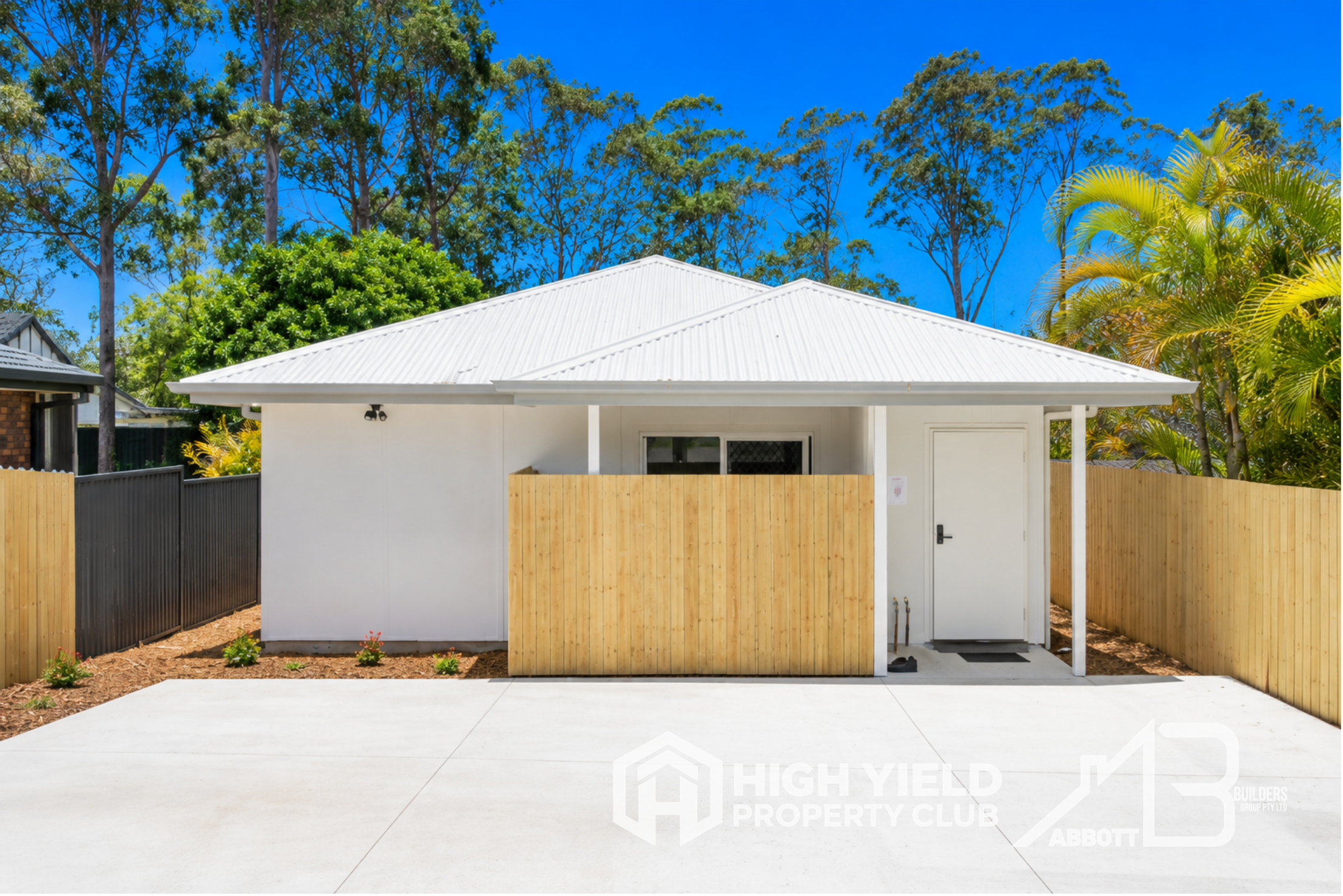 A rooming accomodation house. A small, white, modern house with a gabled metal roof, a wooden fence in front, and a paved area. Tall trees with green foliage and a clear blue sky are in the background.