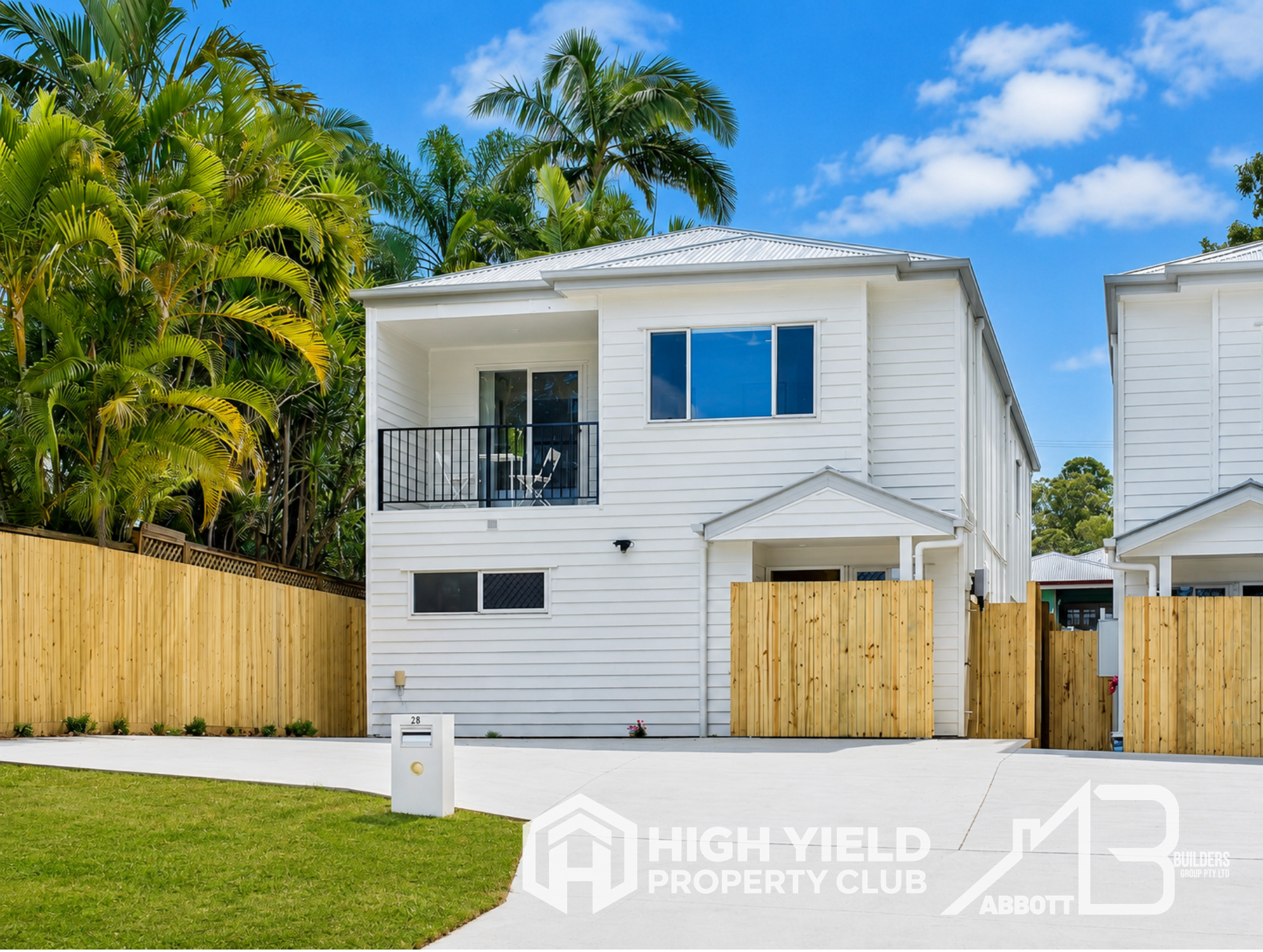 A rooming accomodation house. A white two-story house with a balcony, large windows, and a metallic roof, surrounded by a wooden fence and lush green trees under a blue sky with clouds.