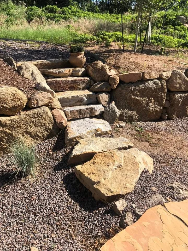Stone steps leading up a small hill with a clay pot on top, surrounded by large rocks and gravel, in a garden or outdoor landscape with green plants and trees in the background.