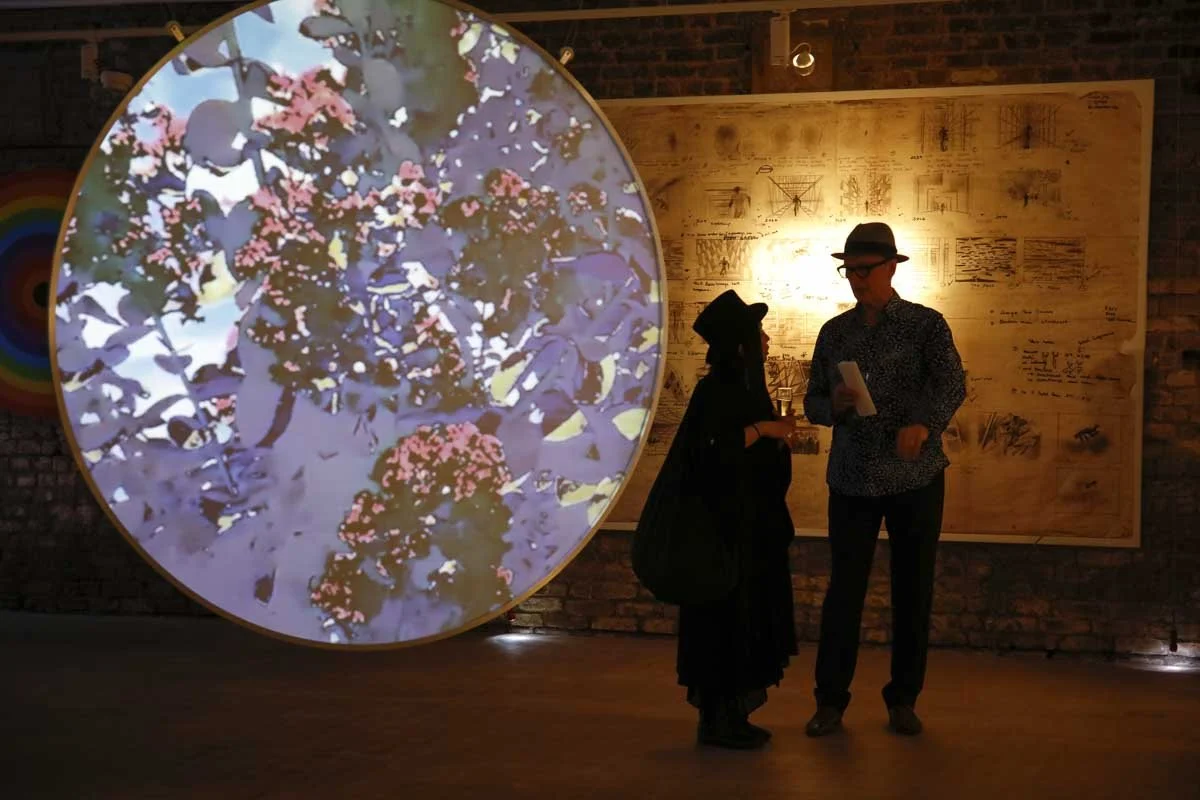 Two people stand and talk in front of a large, illuminated, circular digital art installation of flowers, inside a gallery with exposed brick wall and a large mounted wooden board with sketches and diagrams.Artist Ian Emes