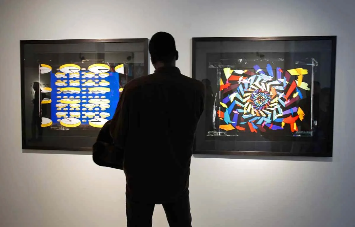 Person standing in front of two framed abstract digital art pieces in a gallery, with one displaying a pattern of yellow and white shapes on a blue background, and the other featuring a colorful, spiraling mosaic of shapes.Artist Ian Emes