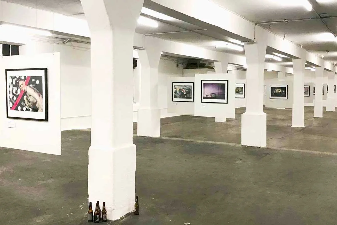 Empty art gallery with white walls, framed photographs on display, support columns, and a few empty bottles on the floor.