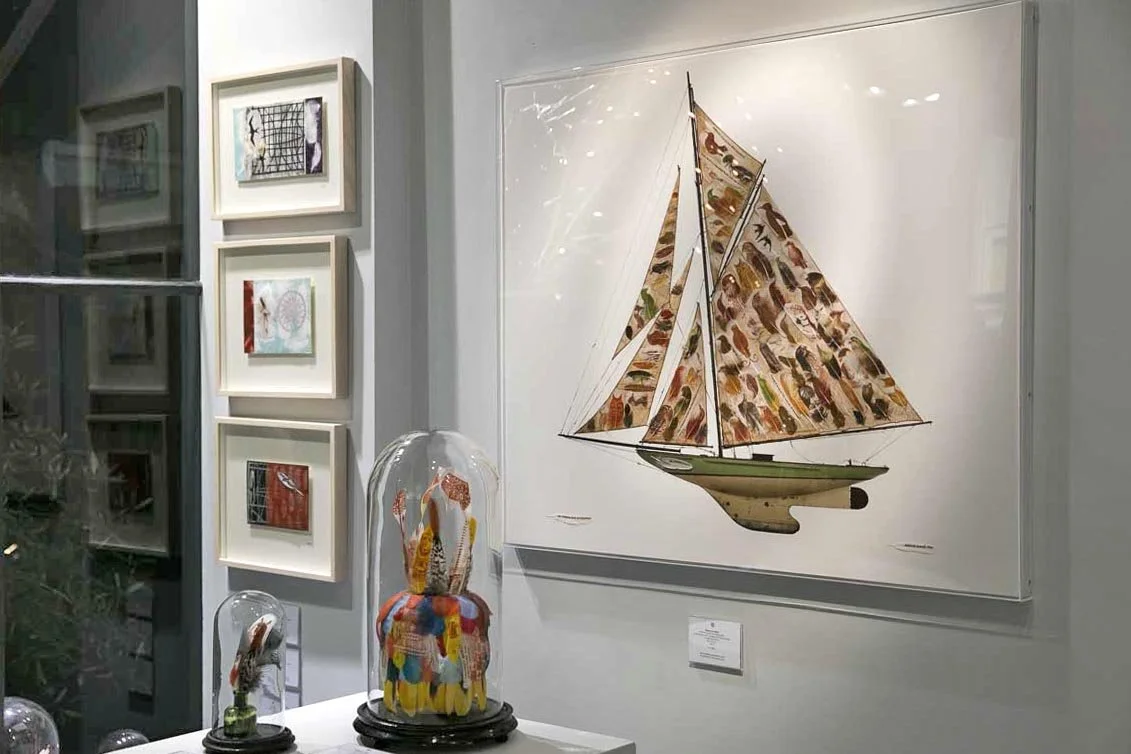 Art gallery display featuring a large framed sailboat painting and several smaller textile artworks under glass domes.