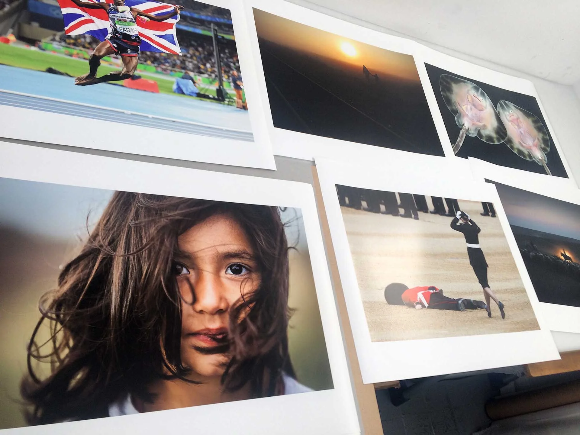 A collage of photographs on a wall. One photo shows a young girl with long curly hair and brown eyes. Other photos include a track athlete holding a British flag at a stadium, a sunset or sunrise scene. Printed for the BPPA assignments exhibition.