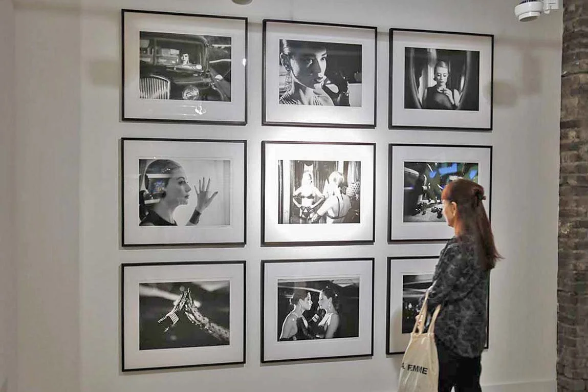 A woman with red hair and a patterned top looks at black-and-white photographs on a gallery wall. Artist Ian Emes