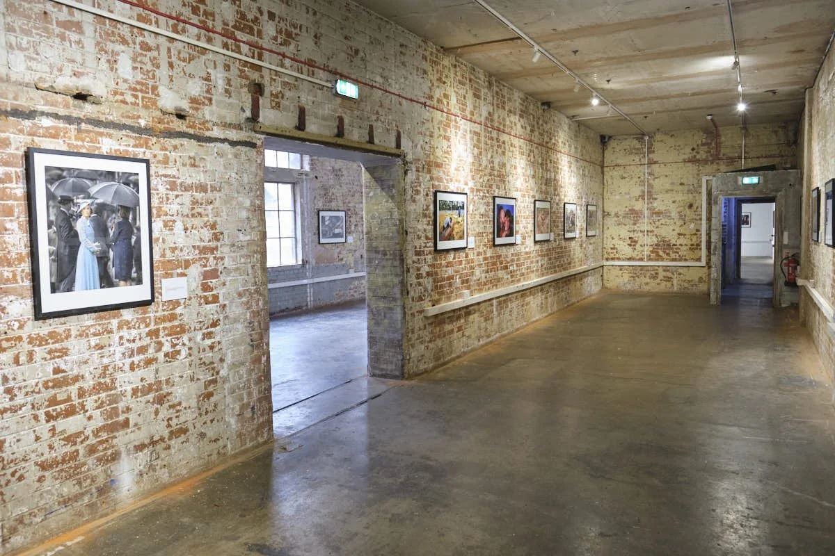 An art gallery with exposed brick walls displaying framed photographs, with a doorway leading to another room with more artwork. The space has a polished concrete floor and overhead lighting.