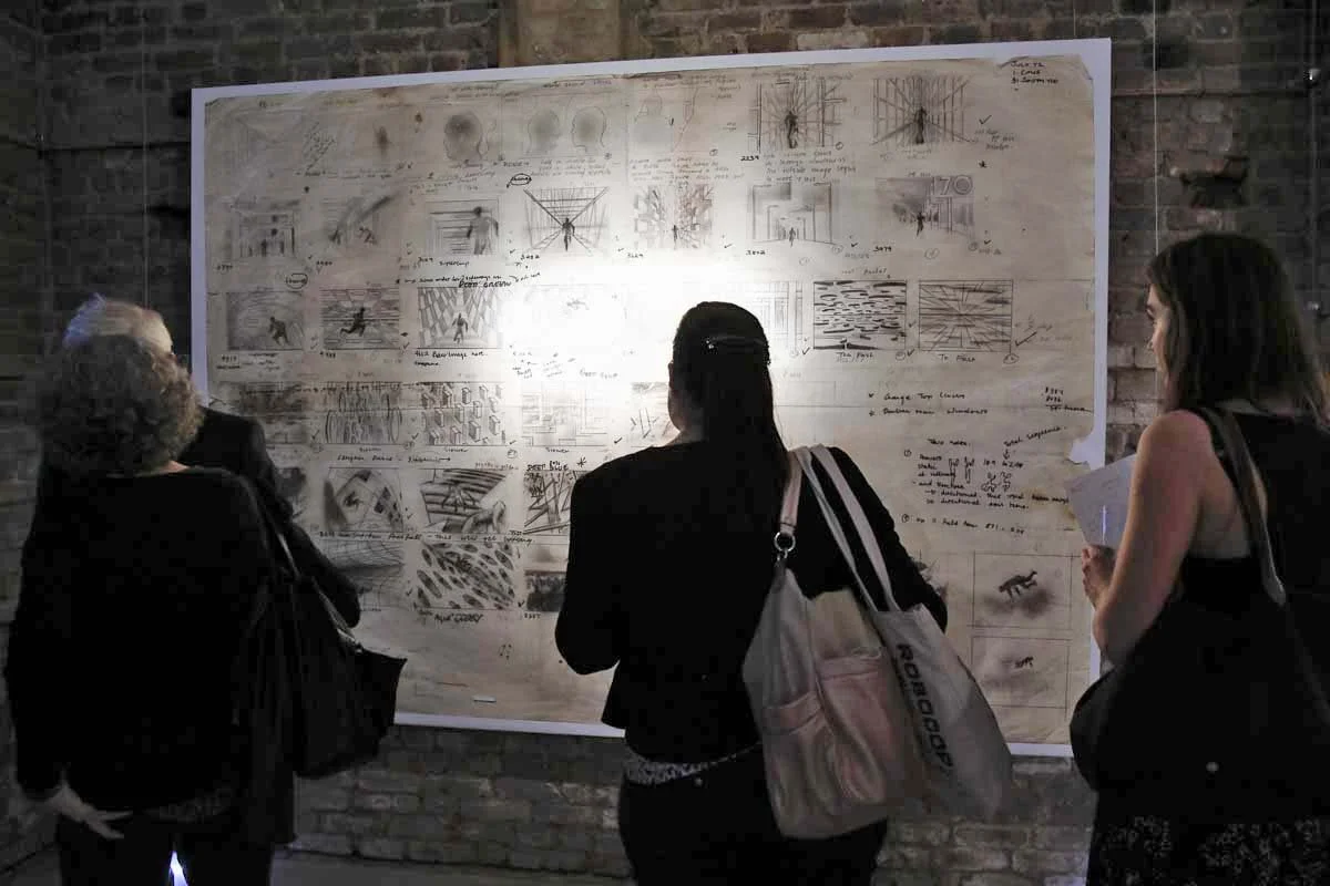 Three people viewing an art display on a large poster board mounted on a brick wall. The display contains sketches, diagrams, and handwritten notes.
