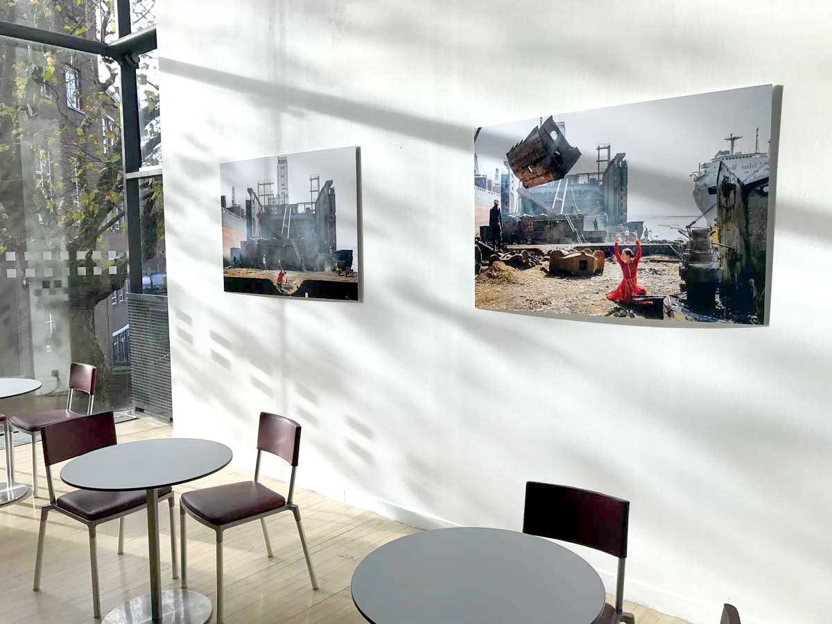 Art gallery interior with two landscape photographs of a shipwreck displayed on a white wall, with sunlight creating shadows. There's a seating area with round tables and chairs near the window. Saddlers Wells London