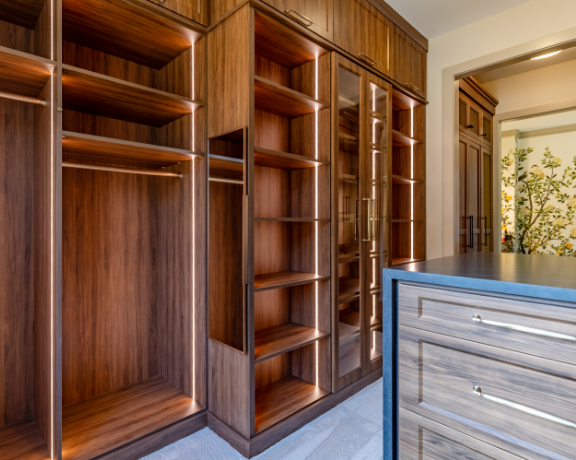 Empty wooden closet with multiple shelves and glass doors, adjacent to a dresser and an open doorway leading to a room with a window and plants.