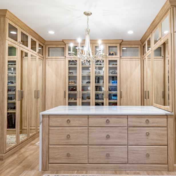A walk-in closet with wooden cabinetry, glass-fronted cabinets, a white marble island, and a chandelier overhead.