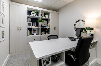 A modern office with a white desk, black office chairs, a bookshelf with decorative items and books, a lamp, and a computer.