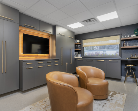 Modern living room with gray cabinets, a wall-mounted TV with backlighting, two tan armchairs, a window with a beige shade, and open shelves with decor items.