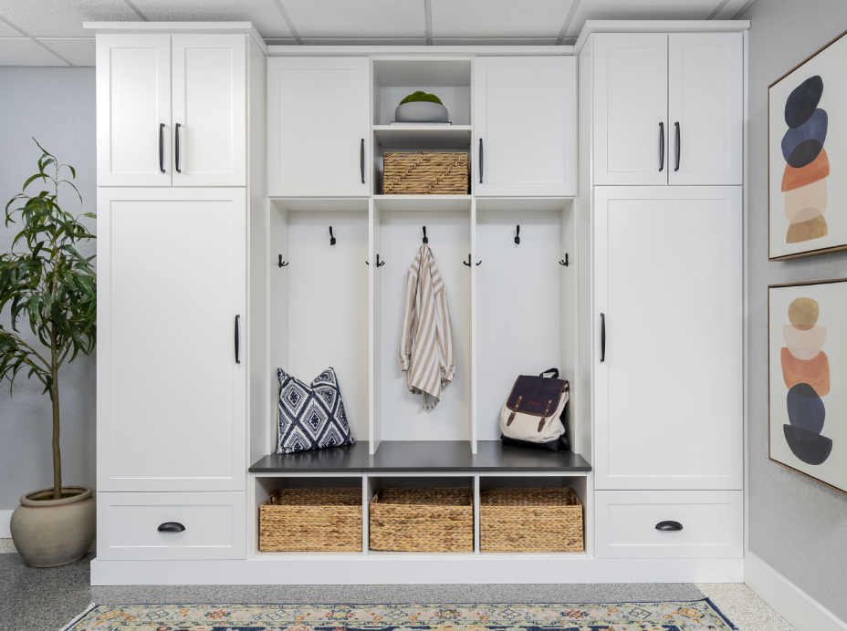 White hallway storage unit with hooks for coats, a bench with baskets underneath, tall cabinets, and decorative art on the wall, with a potted plant on the side.