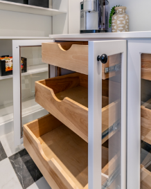 Close-up of a white kitchen cart with three open wooden drawers and a glass panel on the side, placed on a black and white tiled floor.