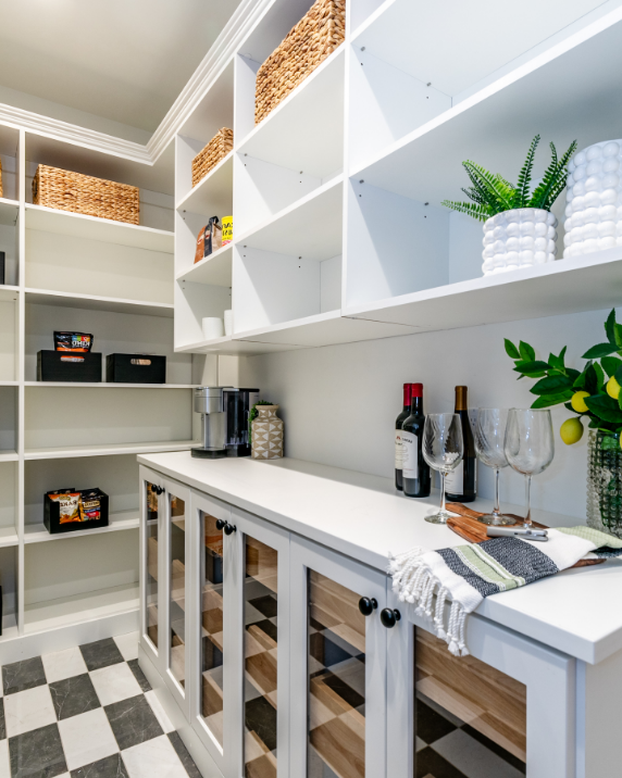 Pantry with white shelves holding woven baskets and snacks, with a white cabinet below containing wine bottles, glasses, and a plant, on a black and white checkered floor.