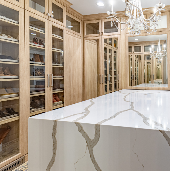 Luxurious walk-in closet with glass-door wooden cabinetry and a marble-topped island, illuminated by chandeliers.