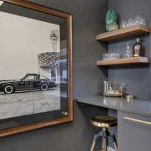 A mirror reflecting a black vintage truck and part of a patterned wall, with a dark gray wall, floating wooden shelves with decorative items, and a small bar or counter with a stool.