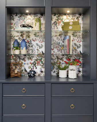 Decorative navy-blue cabinet with glass shelves, containing books, vases, plants, and decorative objects, with floral wallpaper background.