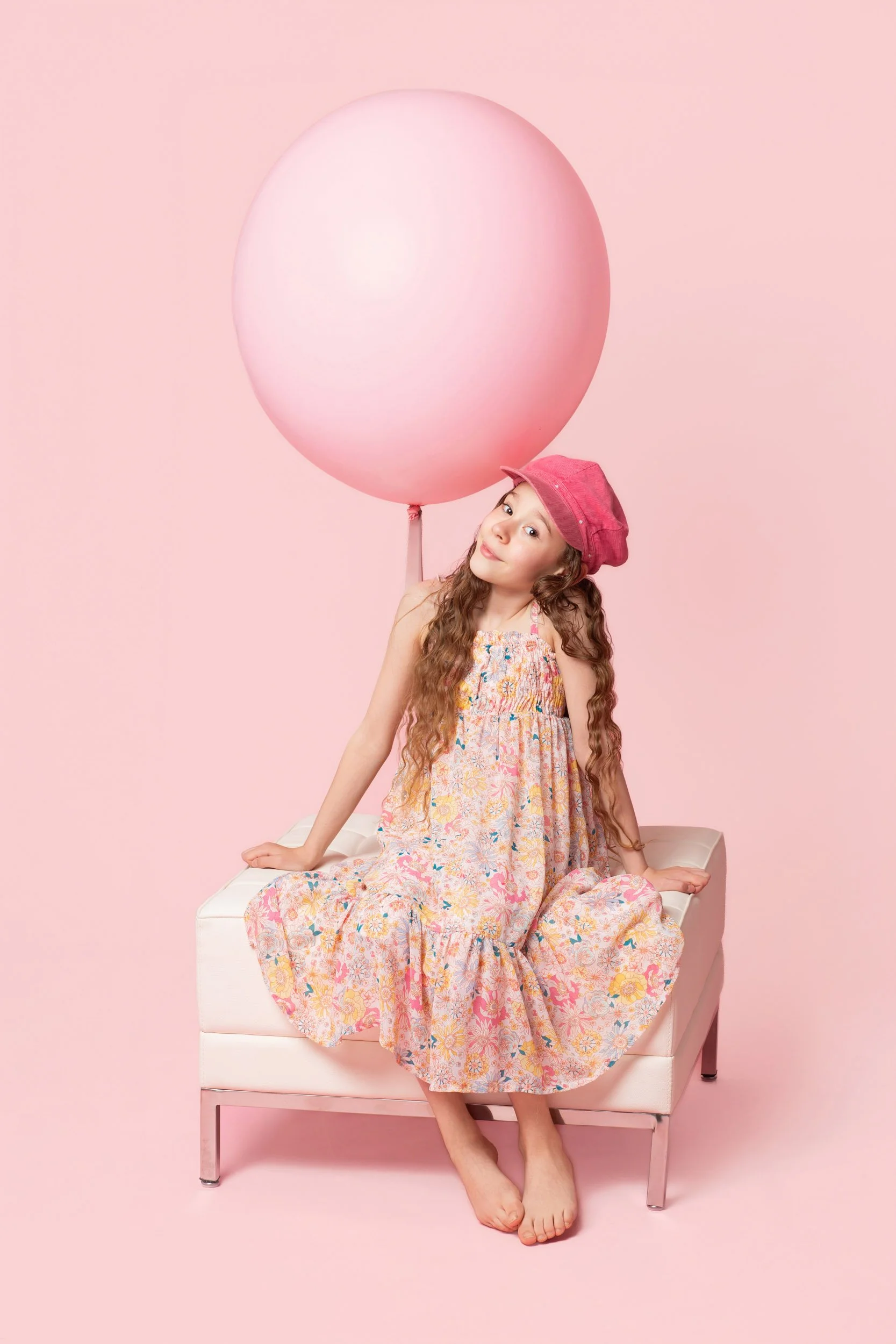 Girl in a floral dress seated on a bench behind an oversized pink balloon during a birthday photoshoot at Poppie Portraits in Seattle.  