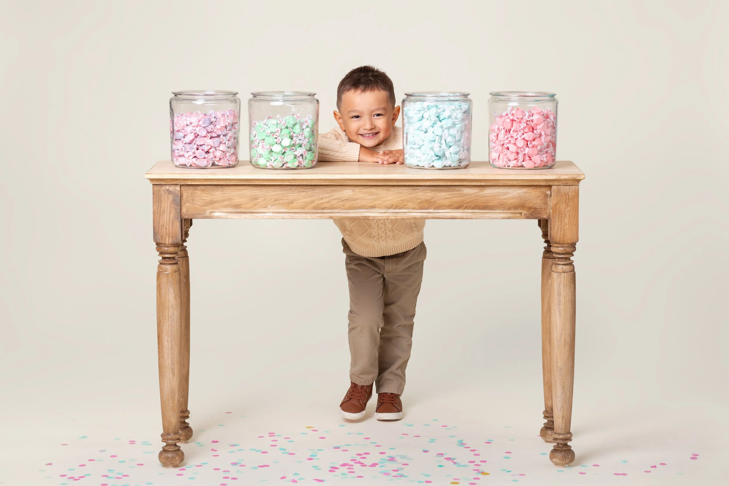 Boy behind a wooden table set with jars of candy on a cream background during a birthday photoshoot at Poppie Portraits in Seattle. 