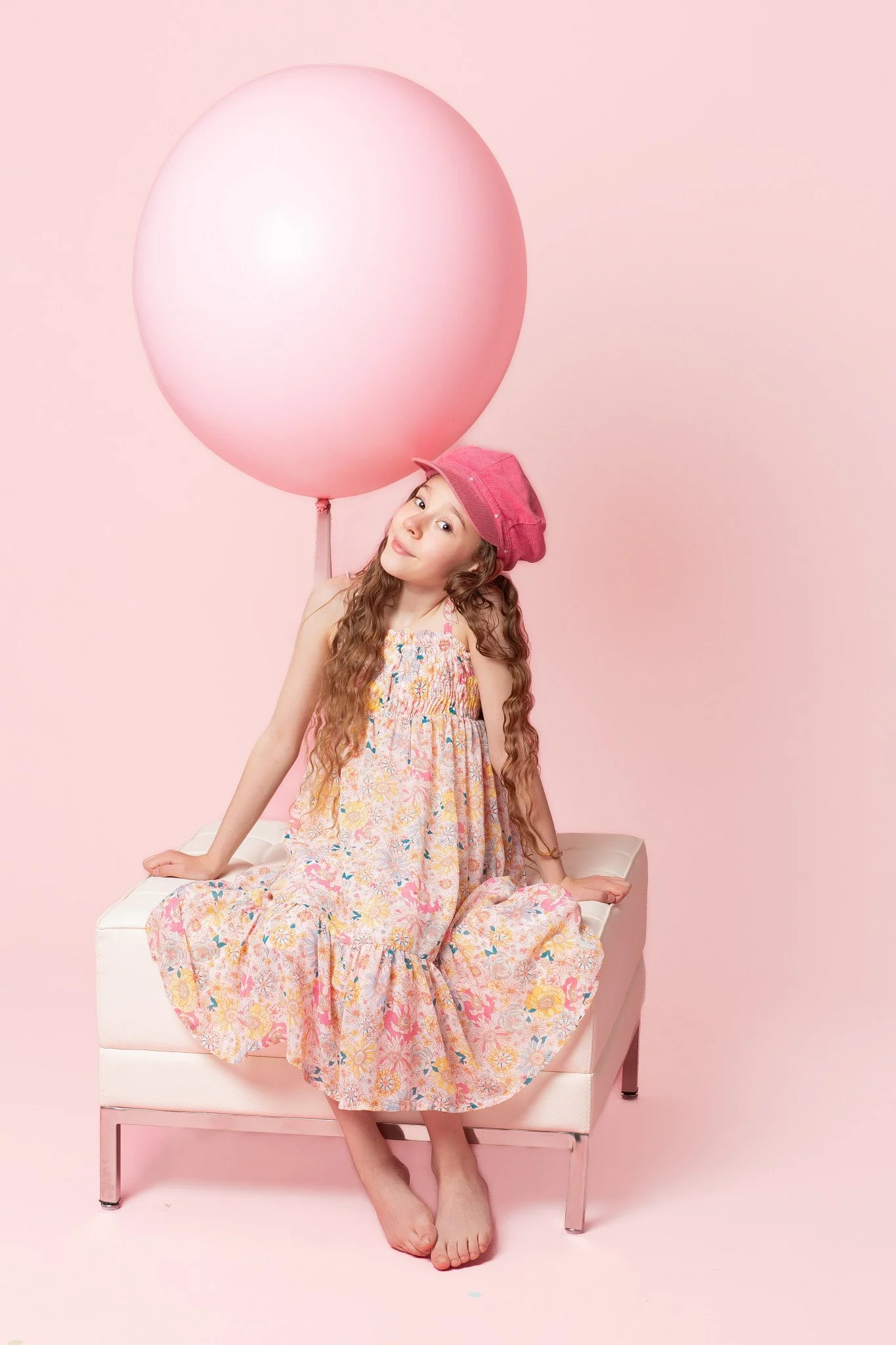 A young girl sitting on a bench on a light coral backdrop holding a jumbo pink balloon during a birthday photoshoot at Poppie Portraits in Seattle.  