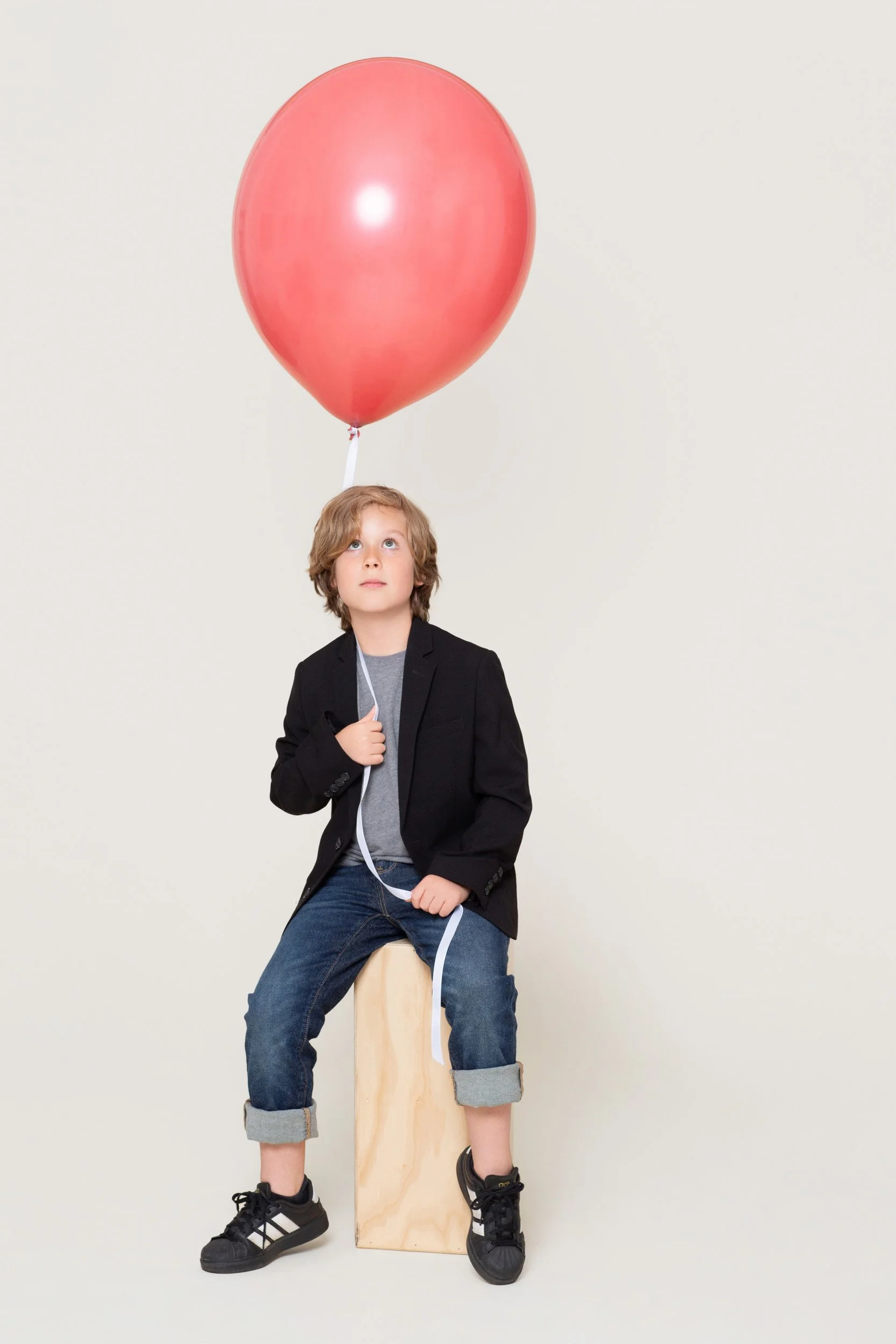 .A smiling boy in a blazer sitting on a wooden bench holding a bouquet of coral balloons during a kids birthday portraits session at Poppie Portraits in Madrona, Seattle.  