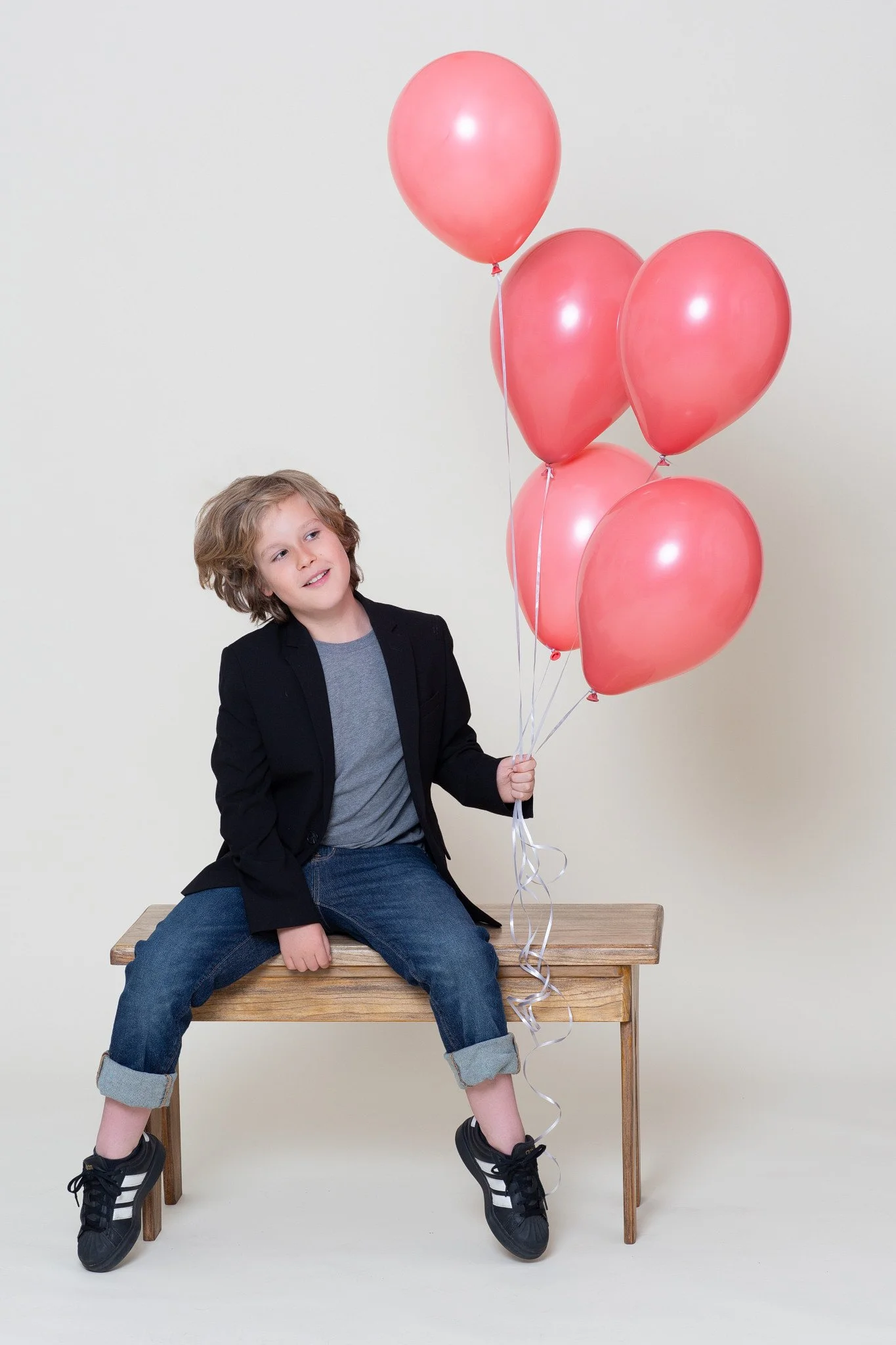 A smiling boy in a blazer sitting on a wooden bench holding a bouquet of coral balloons during a kids birthday portraits session at Poppie Portraits in Madrona, Seattle.  