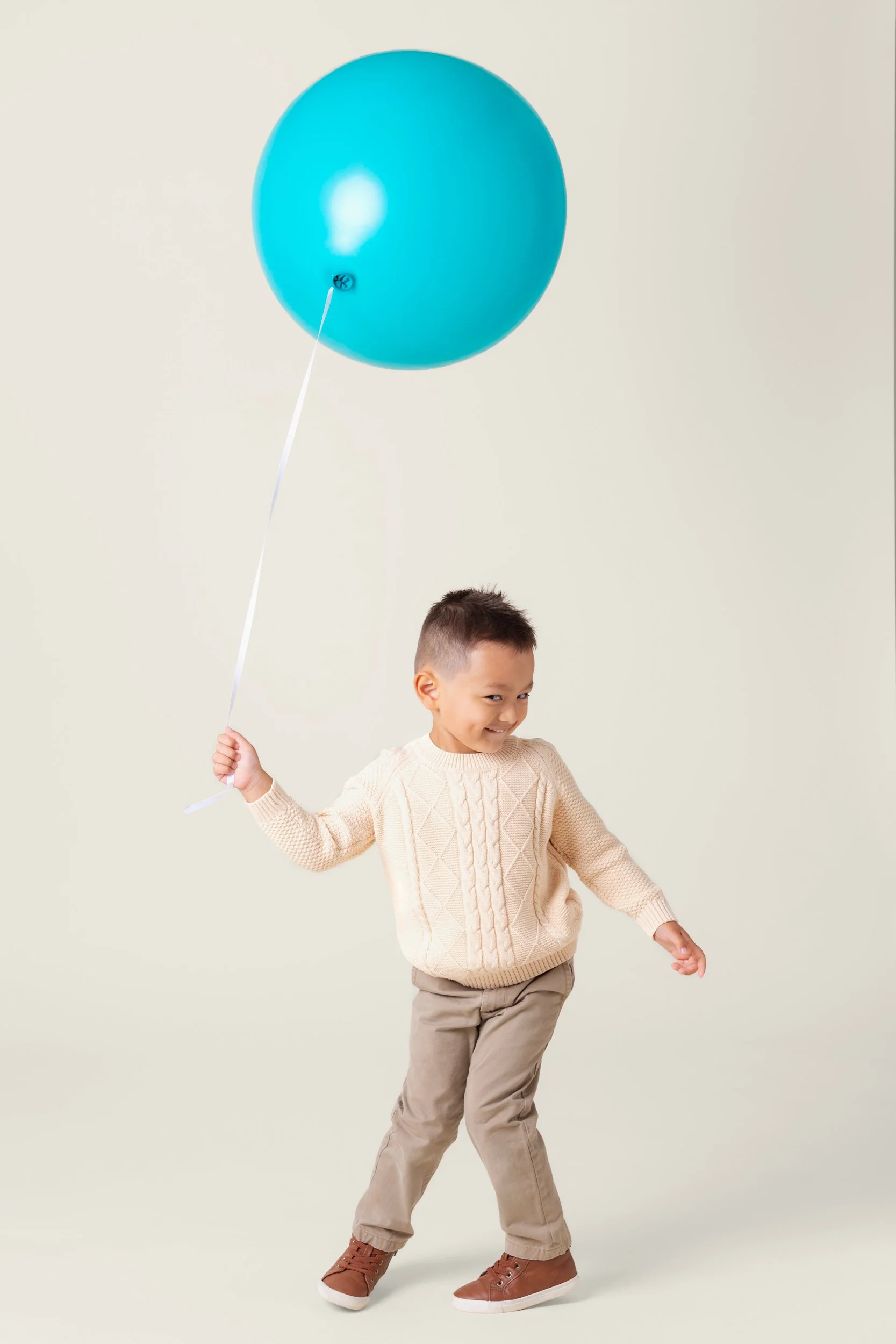 Boy in a cream sweater dancing while holding a turquoise balloon on a string during a birthday photoshoot at Poppie Portraits in Seattle. 