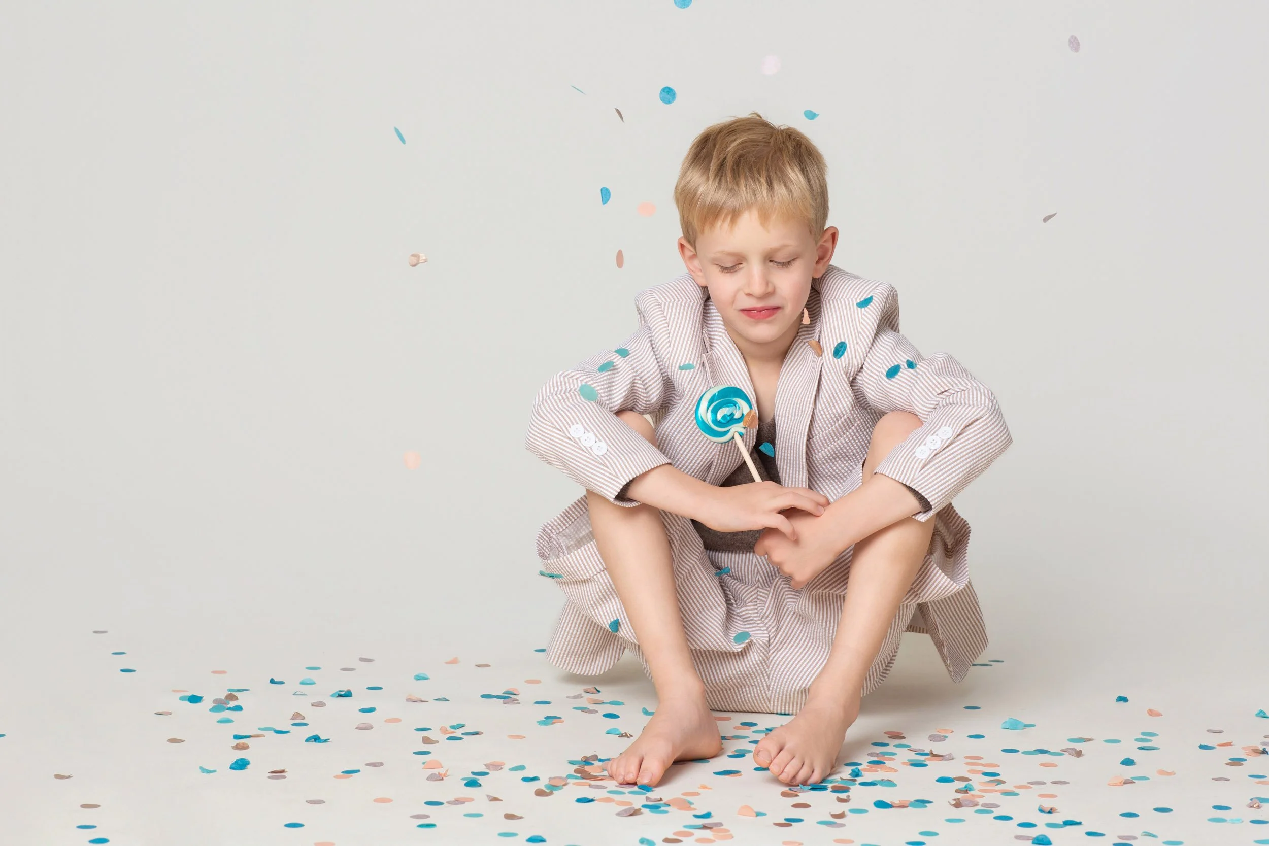 A boy seated on the floor surrounded by scattered confetti with a lollipop during a birthday photoshoot at Poppie Portraits in Seattle. 