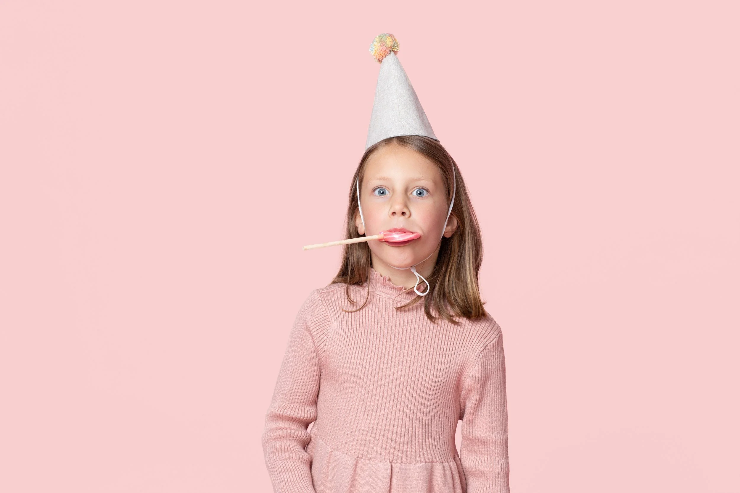 Girl in a pink sweater and party hat on a pink background with a lollipop
