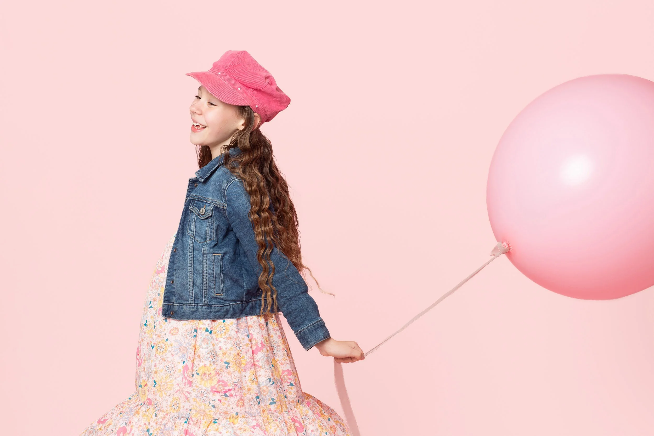 Girl in a pink hat and denim jacket pulling a pink balloon behind her