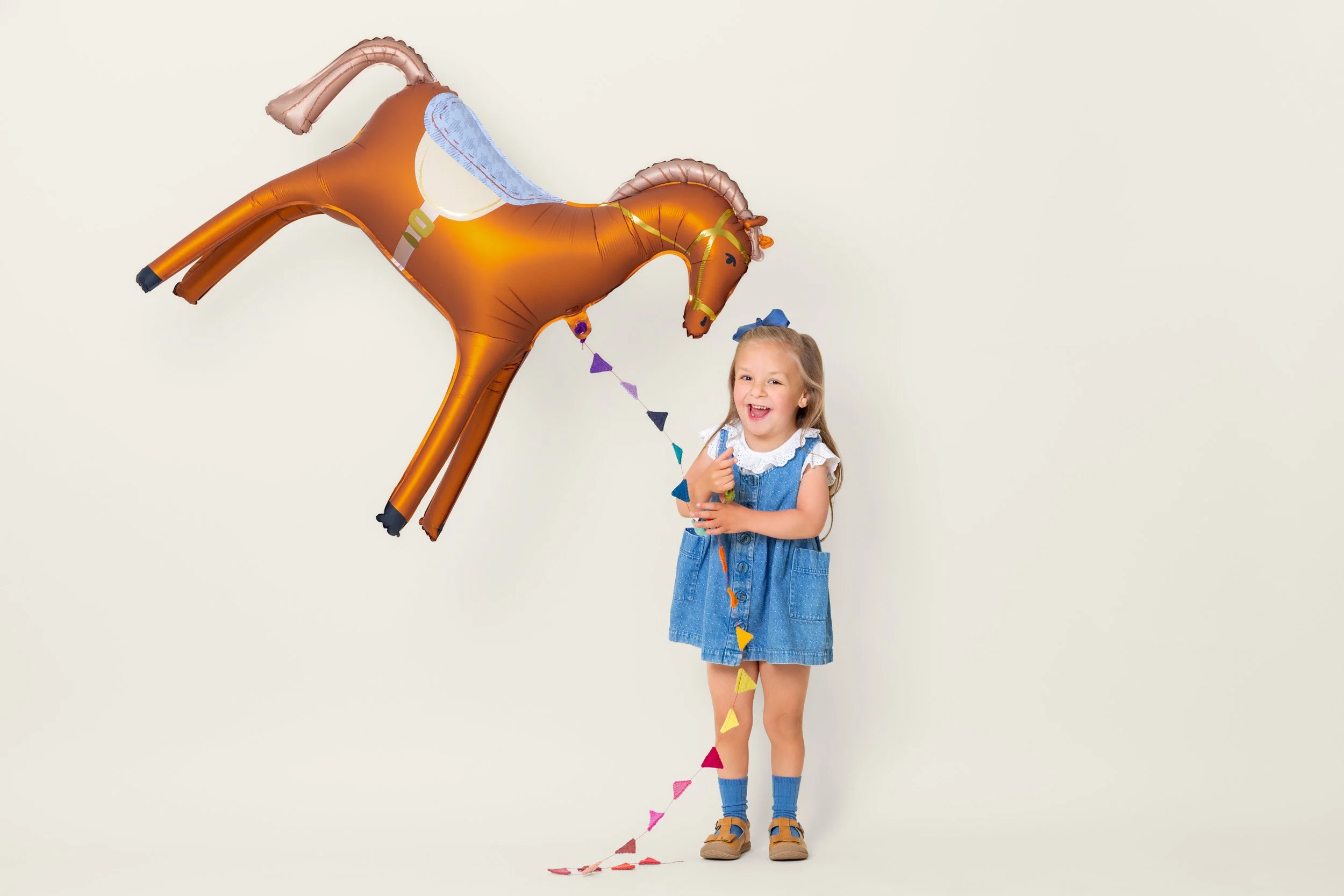 Girl in a blue dress holding the reins of a large horse foil balloon