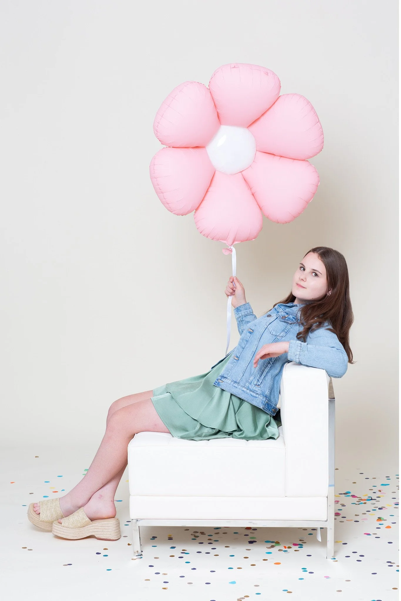 A girl sitting on a miniature white sofa holding a pink flower balloon during a kids birthday portraits session at the Poppie Portraits studio in Madrona, Seattle.  