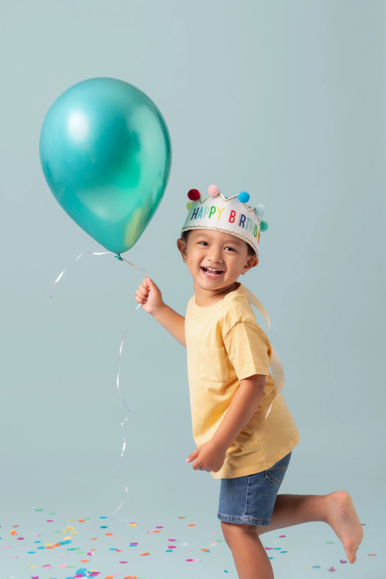 Boy in a yellow shirt and party hat mid-step with a teal balloon and falling confetti during a birthday photoshoot at Poppie Portraits in Seattle. 