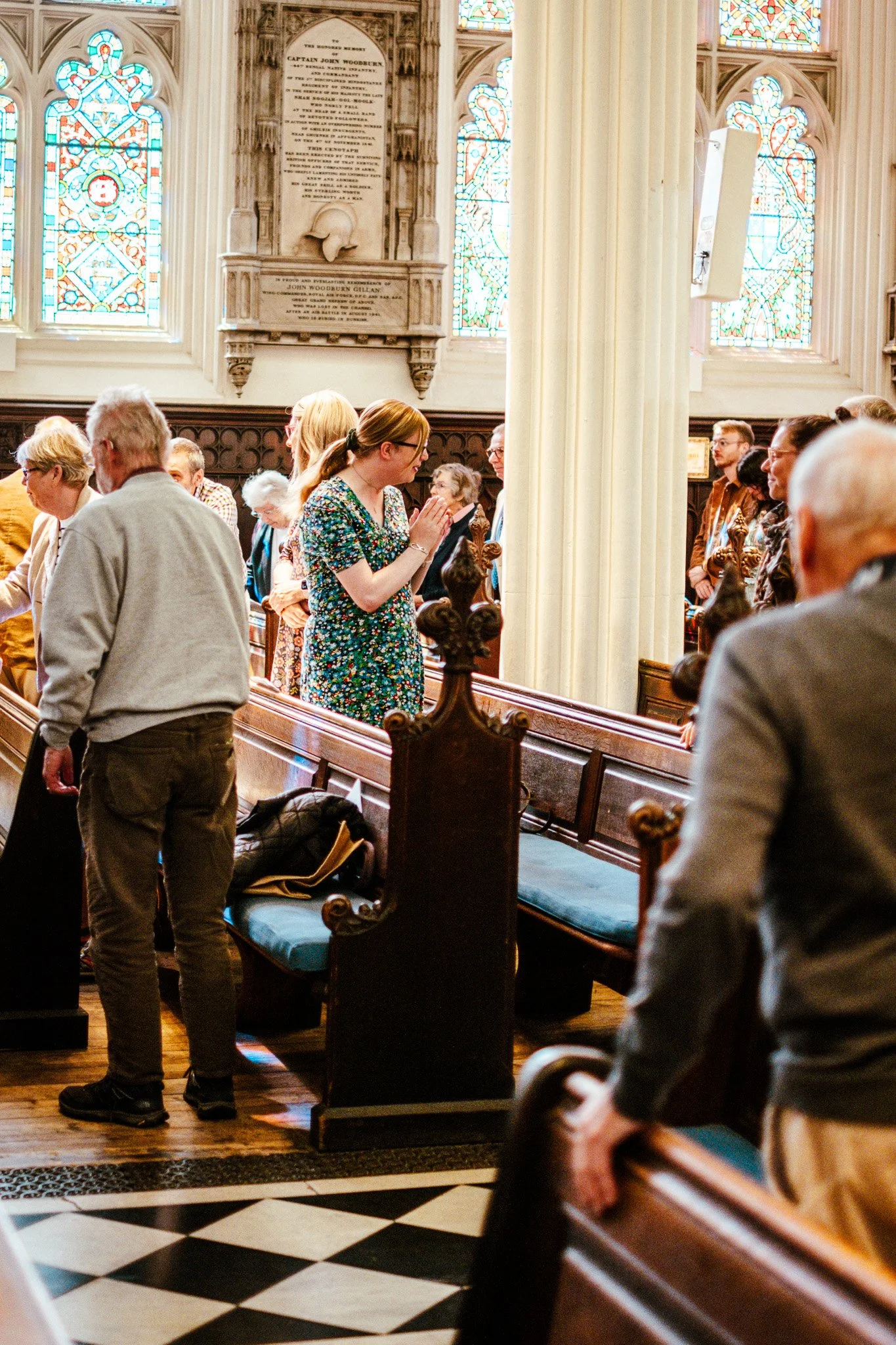 People inside a church or cathedral, standing and sitting in pews, with stained glass windows and religious memorial plaques on the walls.