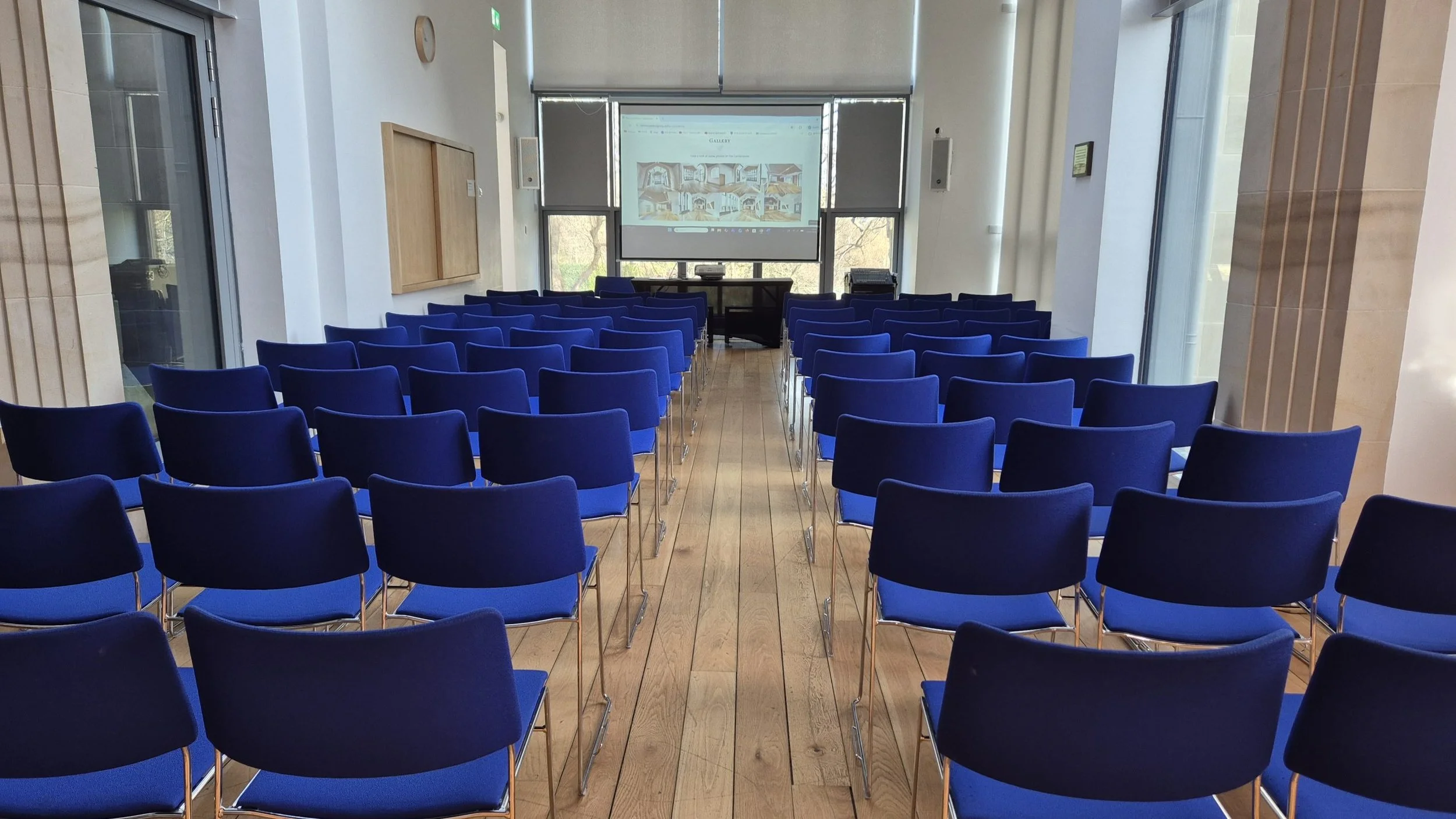 Room set up for a presentation or seminar with rows of blue chairs facing a projection screen displaying a website or presentation slides. Large windows on either side let in natural light.