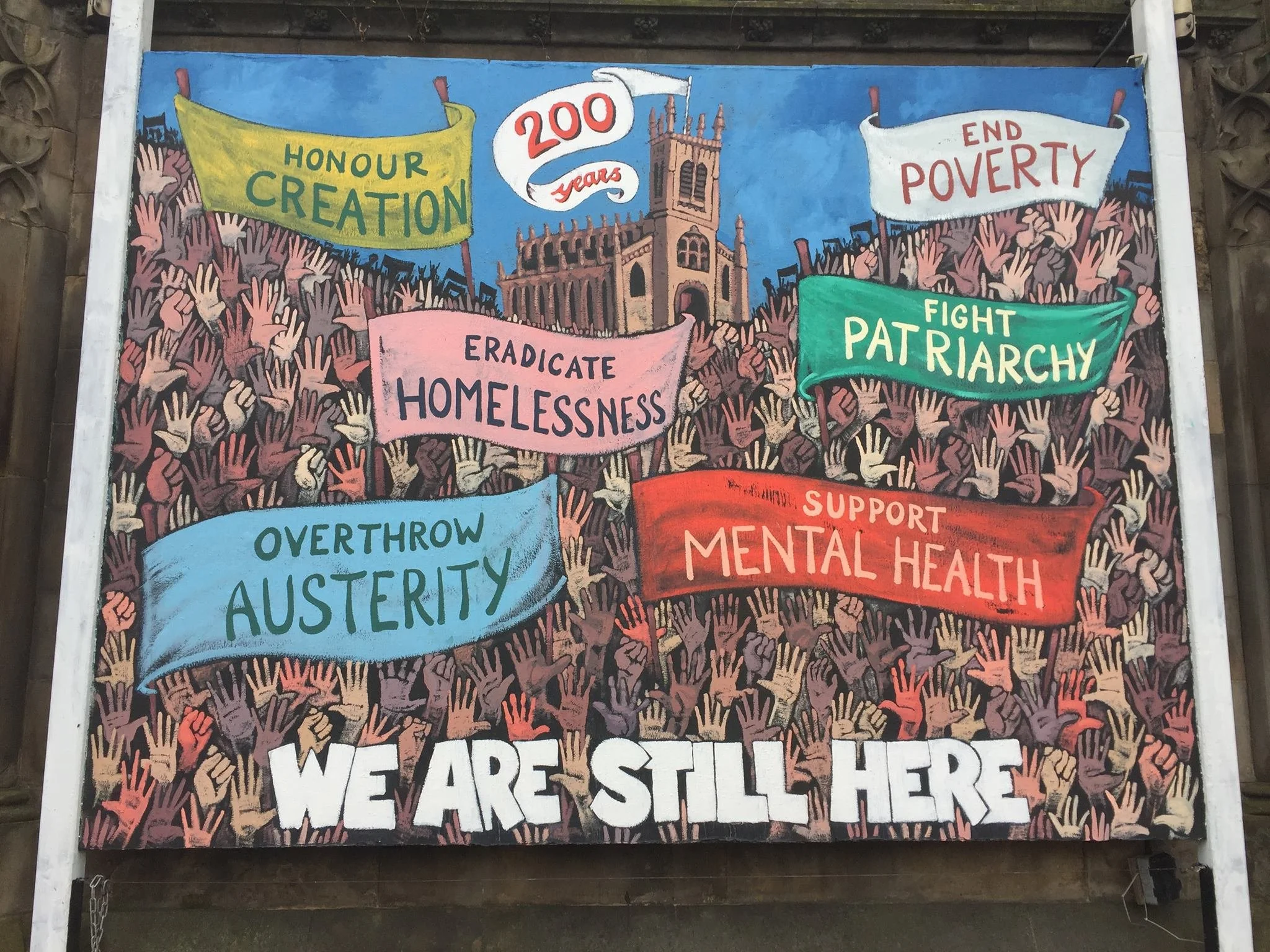 A colorful mural showing a crowd of raised hands with banners advocating for social causes, including honoring creation, ending poverty, eradicating homelessness, fighting patriarchy, supporting mental health, and addressing austerity.