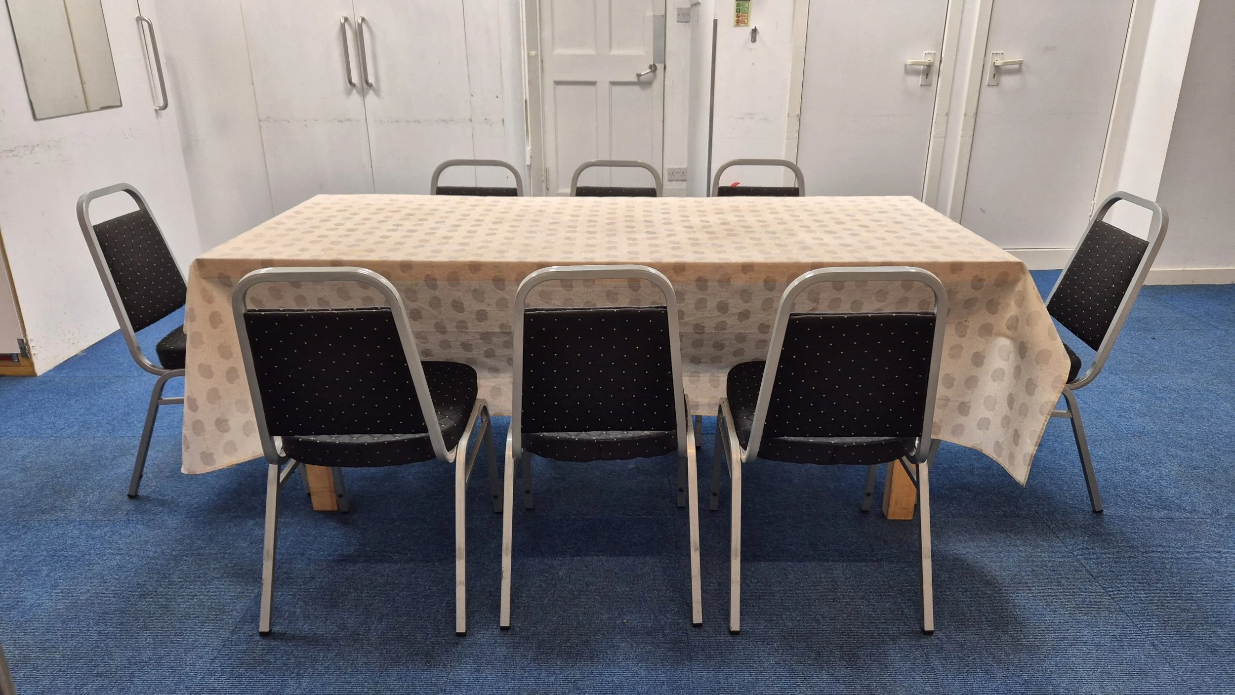 A rectangular table covered with a beige polka dot tablecloth surrounded by six black chairs with silver frames in a room with white walls and blue carpet.