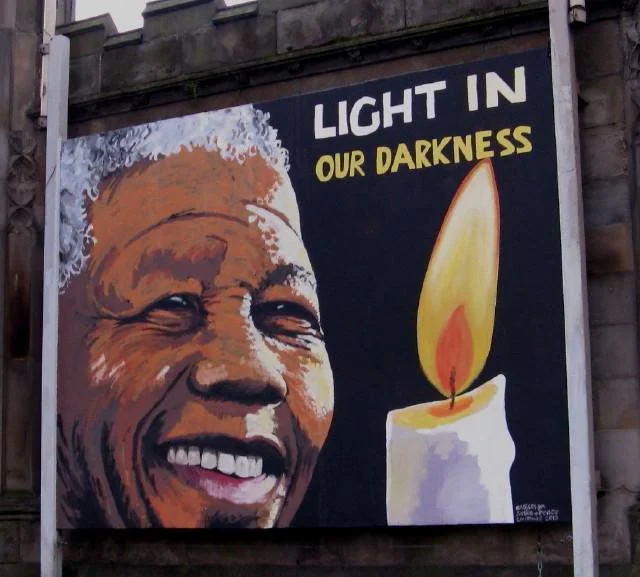 A mural featuring a smiling elderly woman with white hair, alongside a lit candle, and the text 'Light in Our Darkness'.