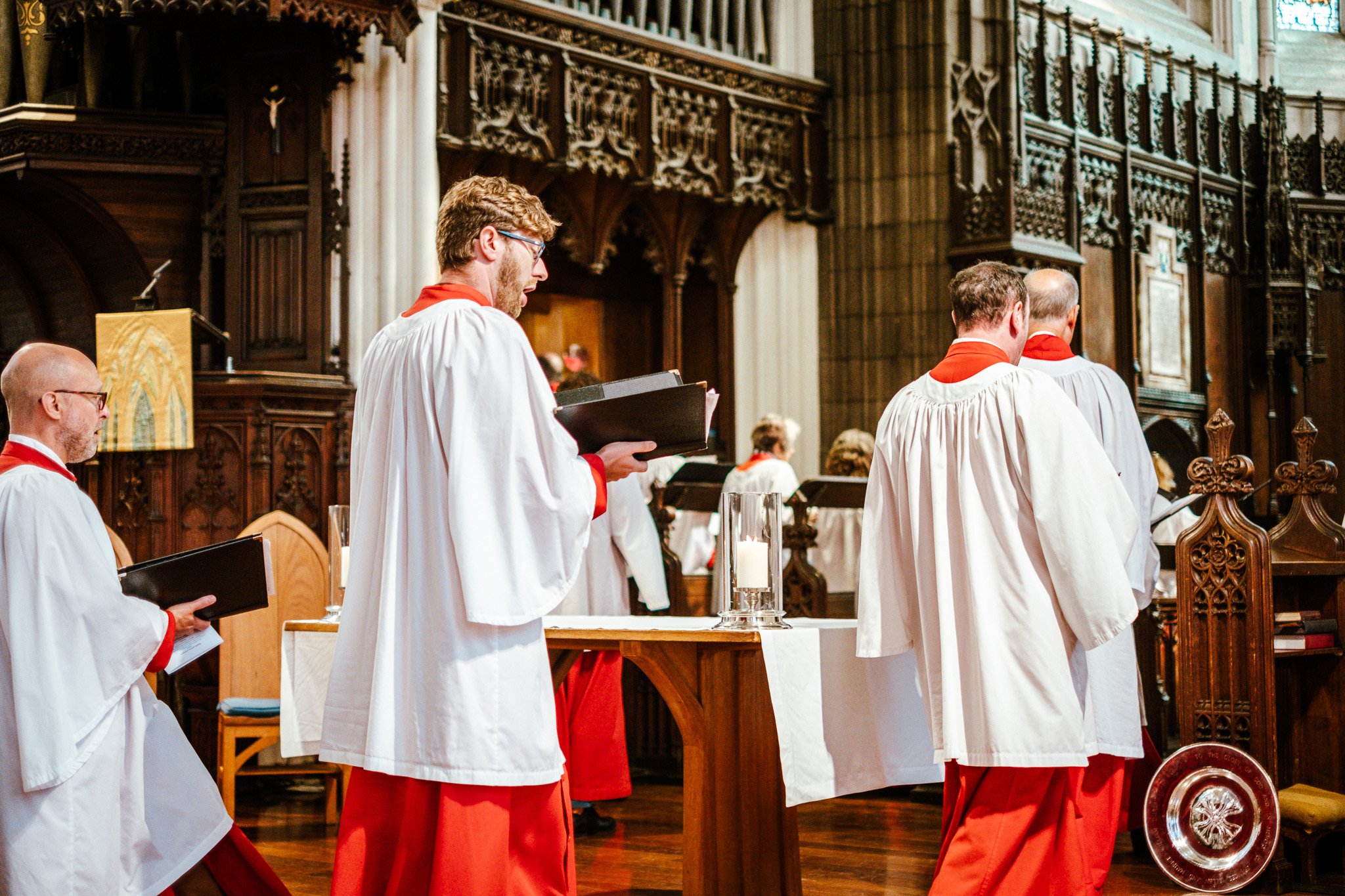 People dressed in church choir robes participating in a religious ceremony inside a church with ornate wooden choir stalls.