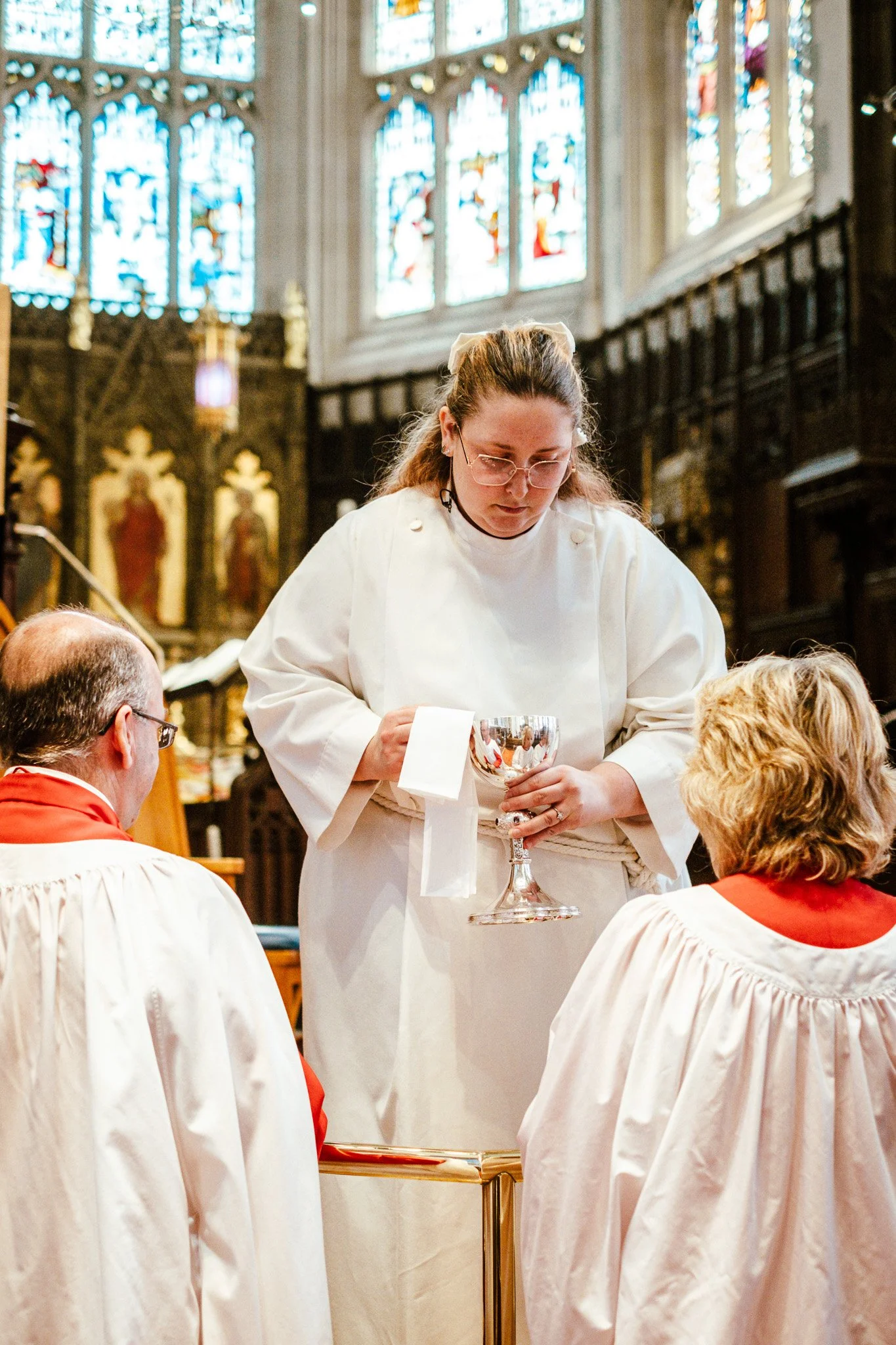 A religious ceremony inside a church with stained glass windows, clergy and congregation members dressed in white and red robes. A young woman is holding a silver chalice.