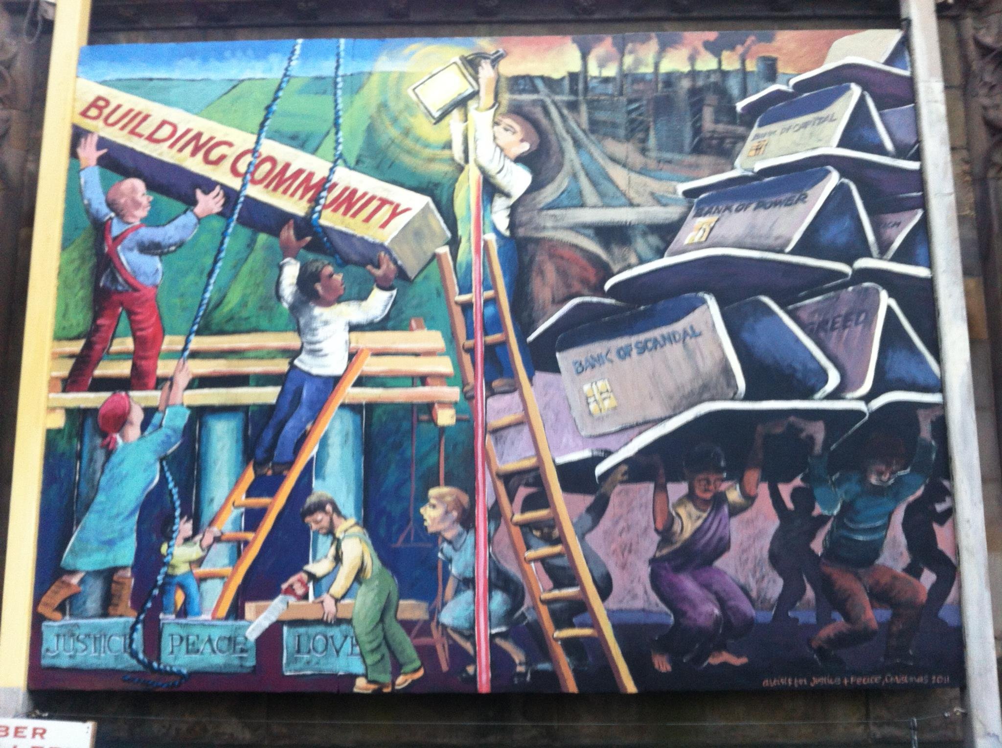 A protest mural depicting people holding a large banner that reads 'BUILDING COMMUNITY' in front of a background showing factory pollution and smokestacks. The mural features children and adults, some climbing ladders and holding signs with words lik