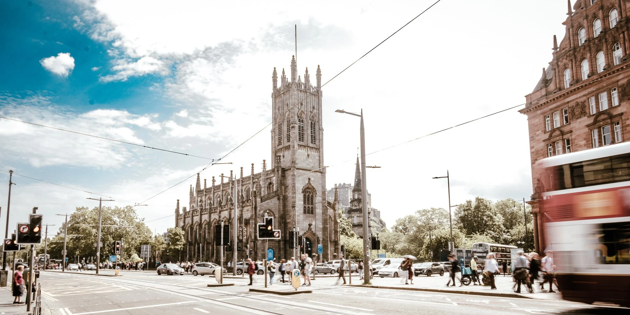A busy city street scene with a historic church tower in the background and a red double-decker bus in motion on the right.