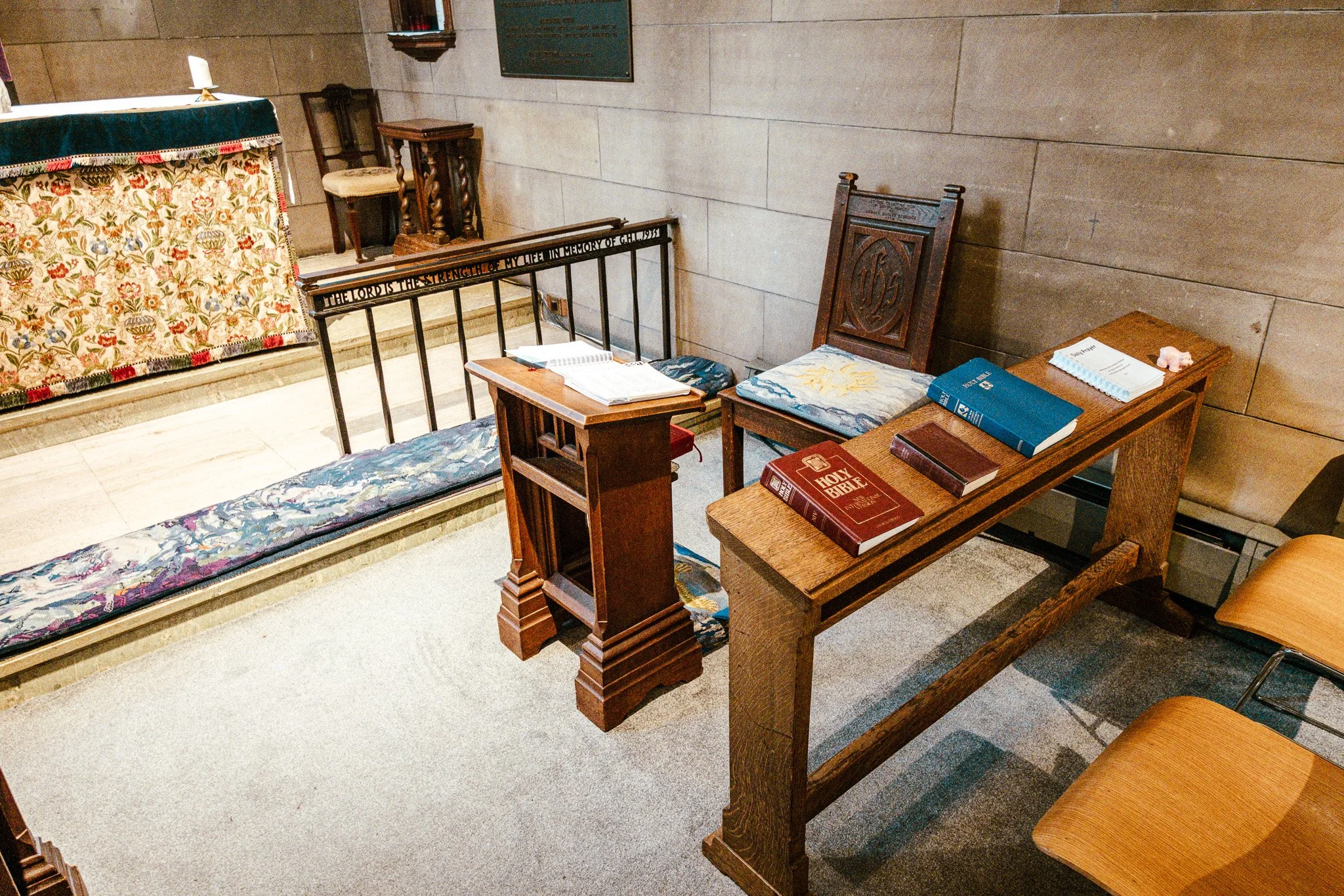 Interior of a church or chapel with wooden chairs, a table holding religious books, and a decorative altar with a floral cloth. The walls are made of stone blocks, and a green plaque or sign is mounted on the wall.