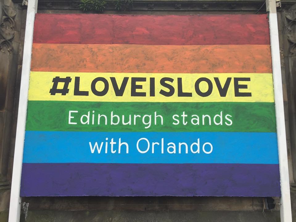 A colorful poster with horizontal stripes in red, orange, yellow, green, blue, and purple, displaying the hashtag #LOVEISLOVE and text about Edinburgh stands with Orlando.