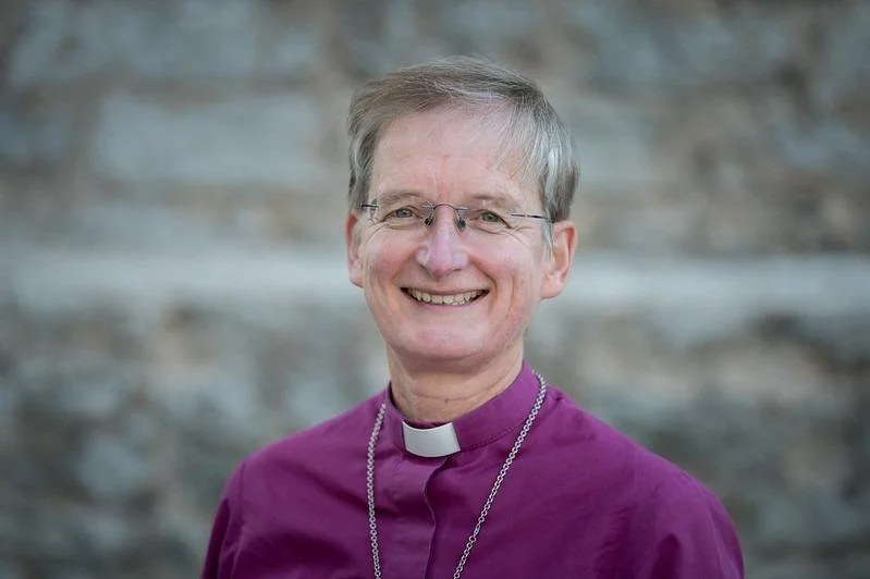 A smiling woman in a purple clerical shirt with a white collar, standing outdoors with a blurred stone wall background.