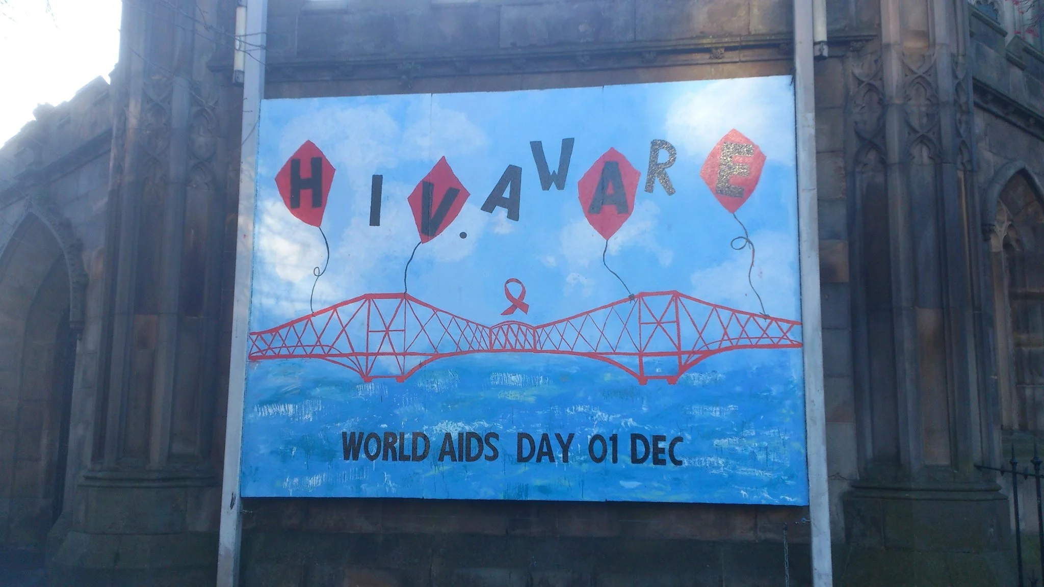 A billboard decoration with the word 'HOPE' spelled out with red diamond-shaped backgrounds and black letters, connected by balloon strings. The billboard features a drawing of a bridge and a red ribbon symbol at the center, with the text 'World AIDS