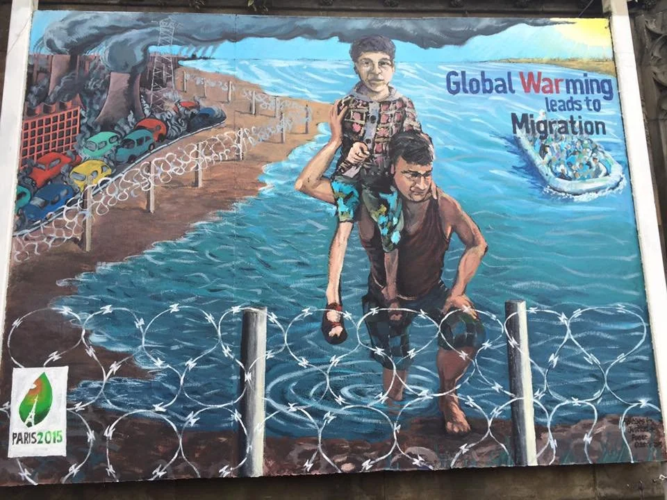 A mural shows a man helping a young boy over a barbed wire fence near a waterway, with a boat and people fleeing in the background. The mural includes the text "Global Warming leads to Migration" and the Paris 2015 logo.